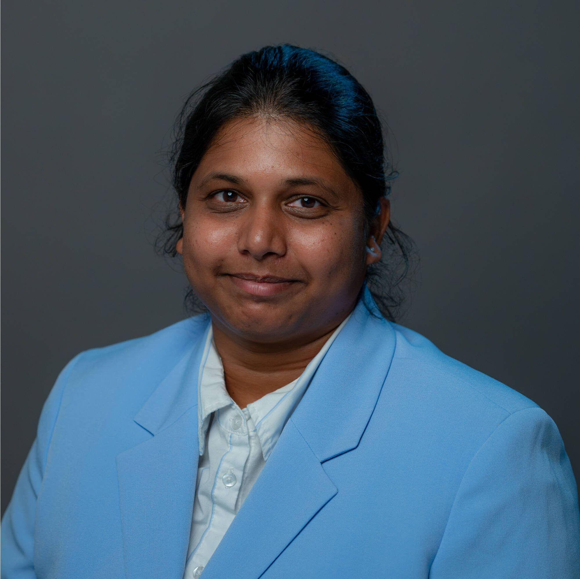 Professional headshot of a Tejaswi Manchineella smiling, wearing a light blue blazer over a white shirt against a dark gray background.